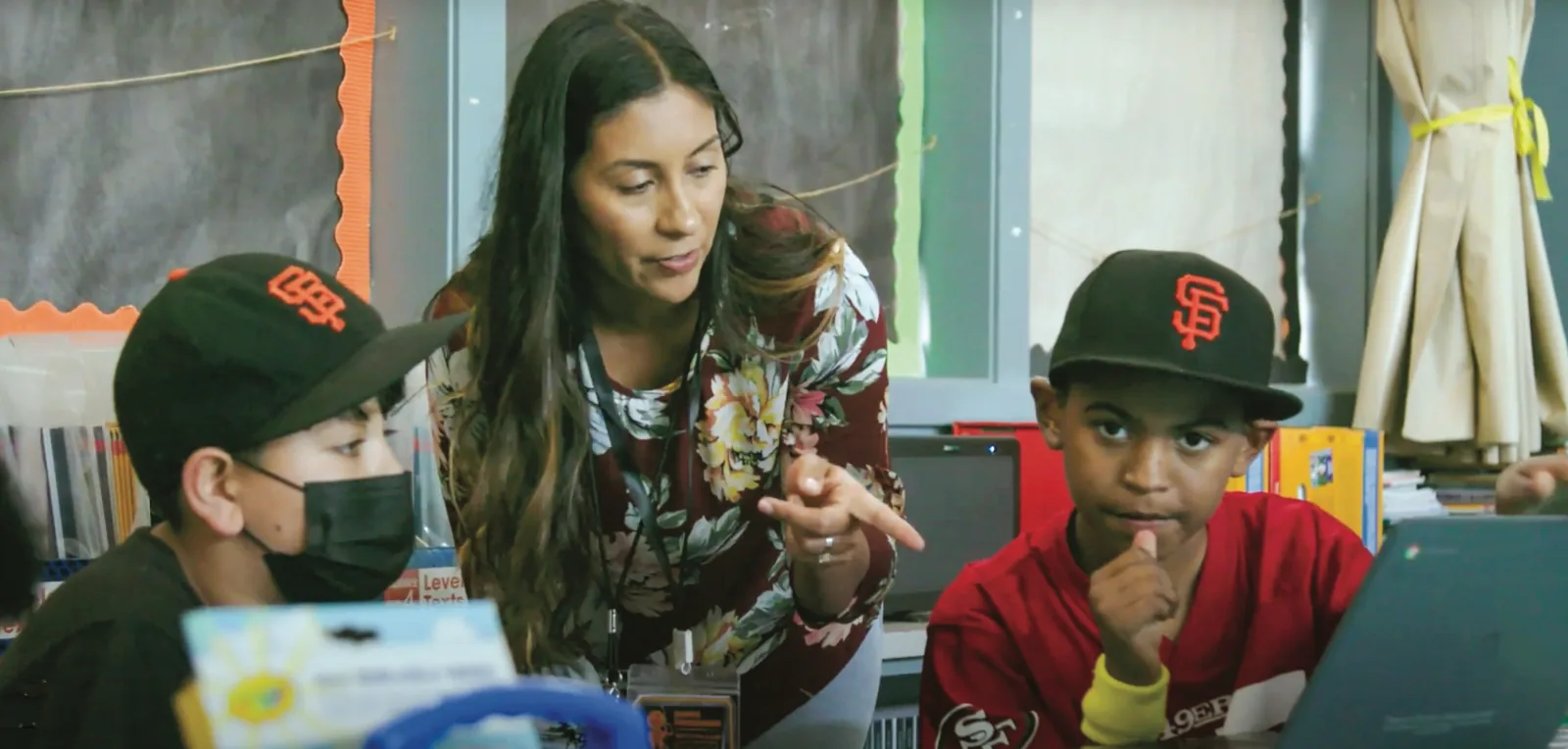A teacher talks with two students at a desk with an open laptop